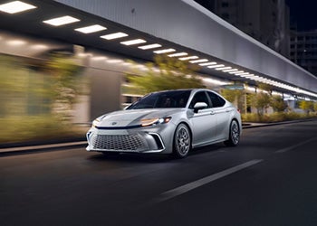 AToyota Camry Hybrid driving through a city street illuminated by streetlights at night.